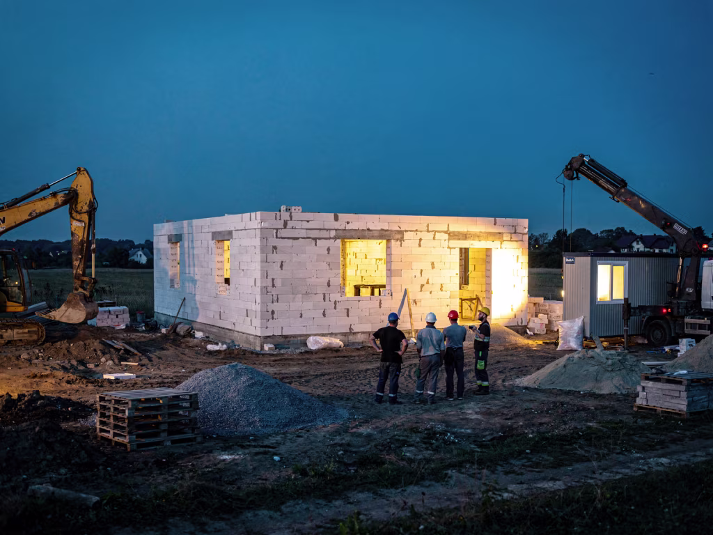 Будівельники в касках біля коробки будинку з газоблоку ввечері, поруч екскаватор і кран